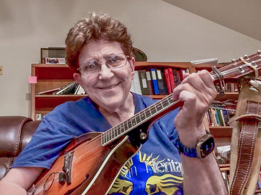 Suzy Oliver, a woman in her 60's with short brown curly hair, beaming a very happy smile, wearing a navy fiddling contest t-shirt, and holding her mandolin. Her bookcases filled with books are in the background.